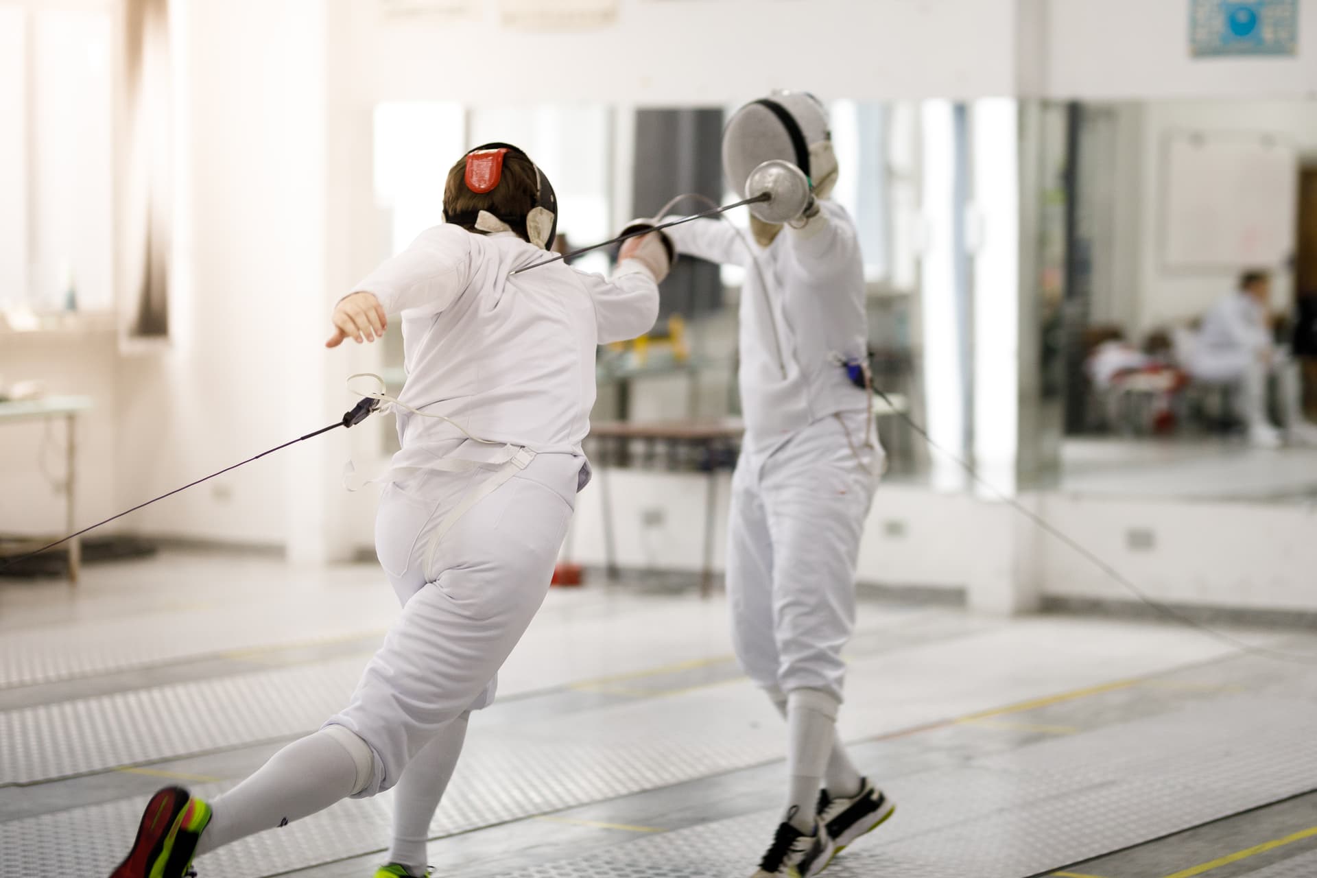 Two fencers on a metal piste, one is fleching, the other is hitting them on the back